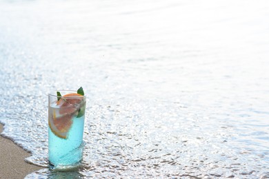 Photo of Glass of refreshing drink with grapefruit and mint getting hit by wave on sea beach, space for text