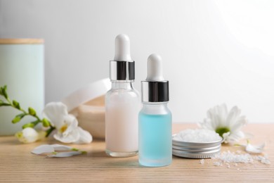 Photo of Bottles of cosmetic serum, sea salt and beautiful flowers on wooden table, closeup