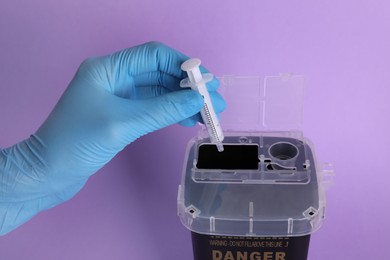 Doctor throwing used syringe into sharps container on violet background, closeup