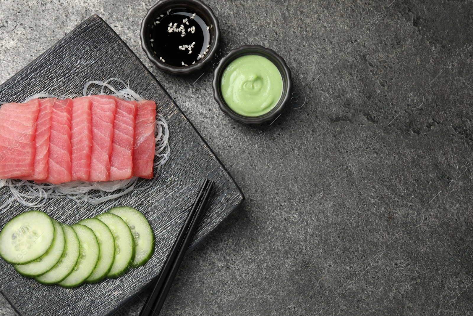 Photo of Tasty sashimi (pieces of fresh raw tuna), cucumber slices, glass noodles, soy sauce and wasabi on gray table, flat lay. Space for text