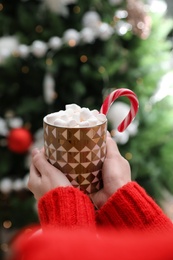 Photo of Woman with cup of delicious hot drink near Christmas tree indoors, closeup