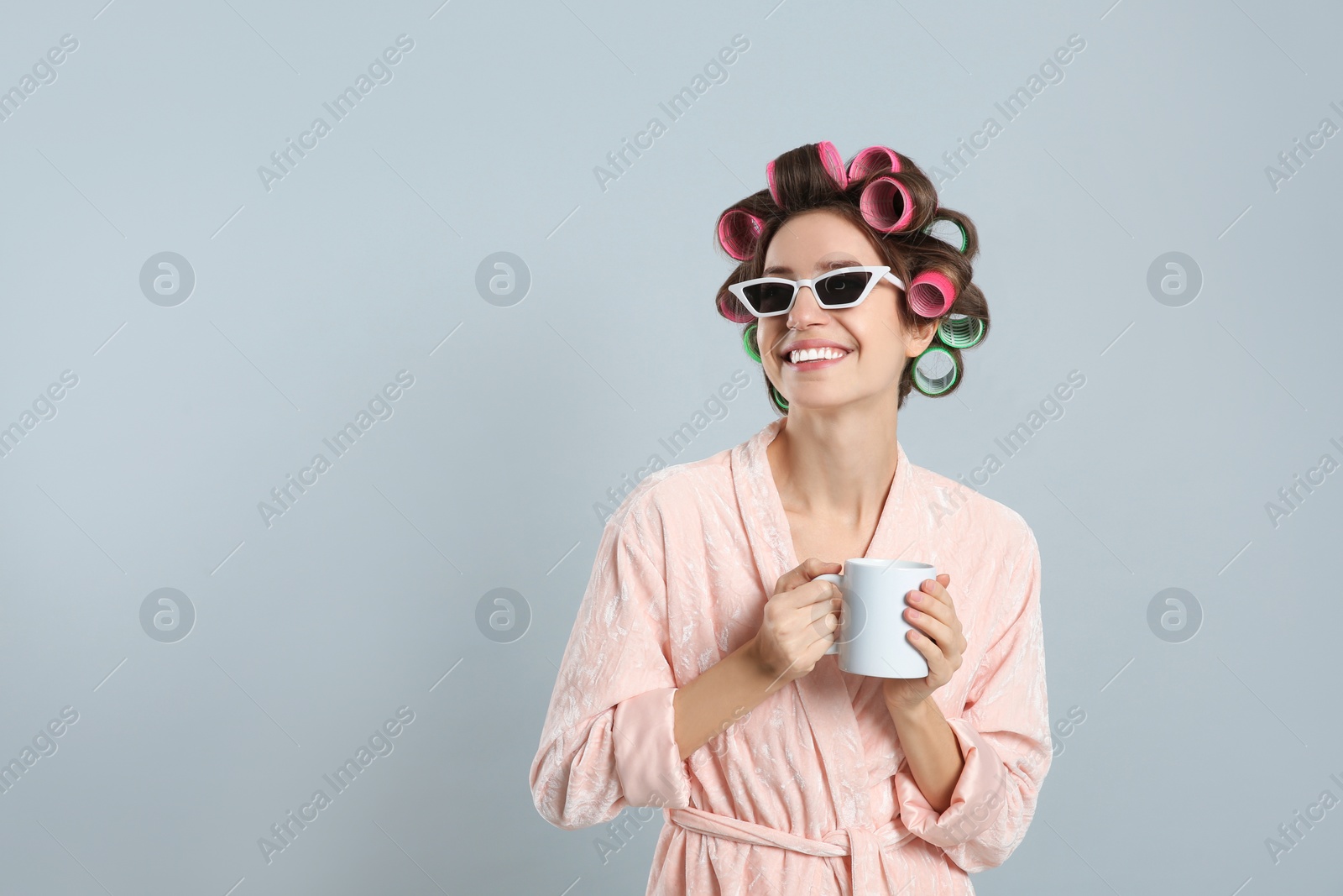 Photo of Beautiful young woman in bathrobe with hair curlers holding cup of drink on light grey background. Space for text