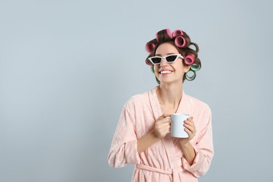 Photo of Beautiful young woman in bathrobe with hair curlers holding cup of drink on light grey background. Space for text