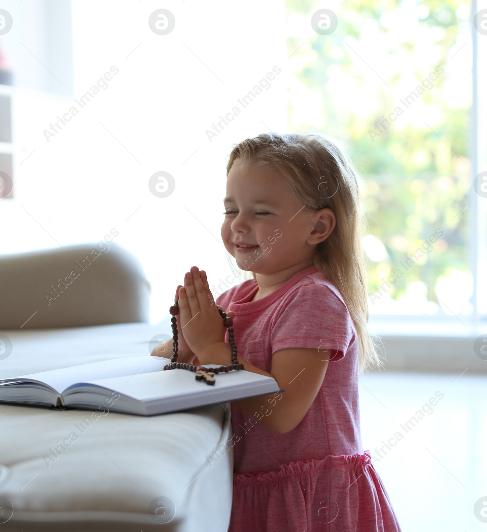 Photo of Cute little girl with beads praying over Bible in living room