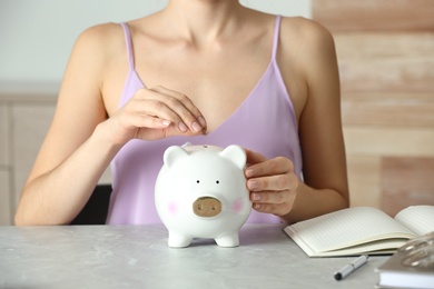 Photo of Woman putting coin into piggy bank at grey marble table, closeup. Money savings