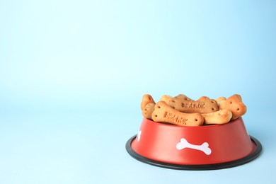 Bone shaped dog cookies in feeding bowl on light blue background, space for text
