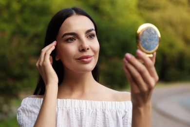 Beautiful young woman looking at herself in cosmetic pocket mirror outdoors