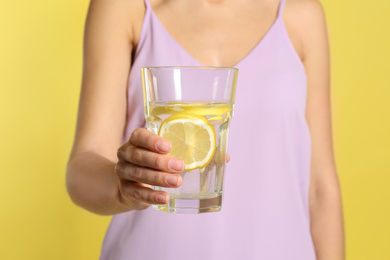 Young woman with glass of lemon water on yellow background, closeup