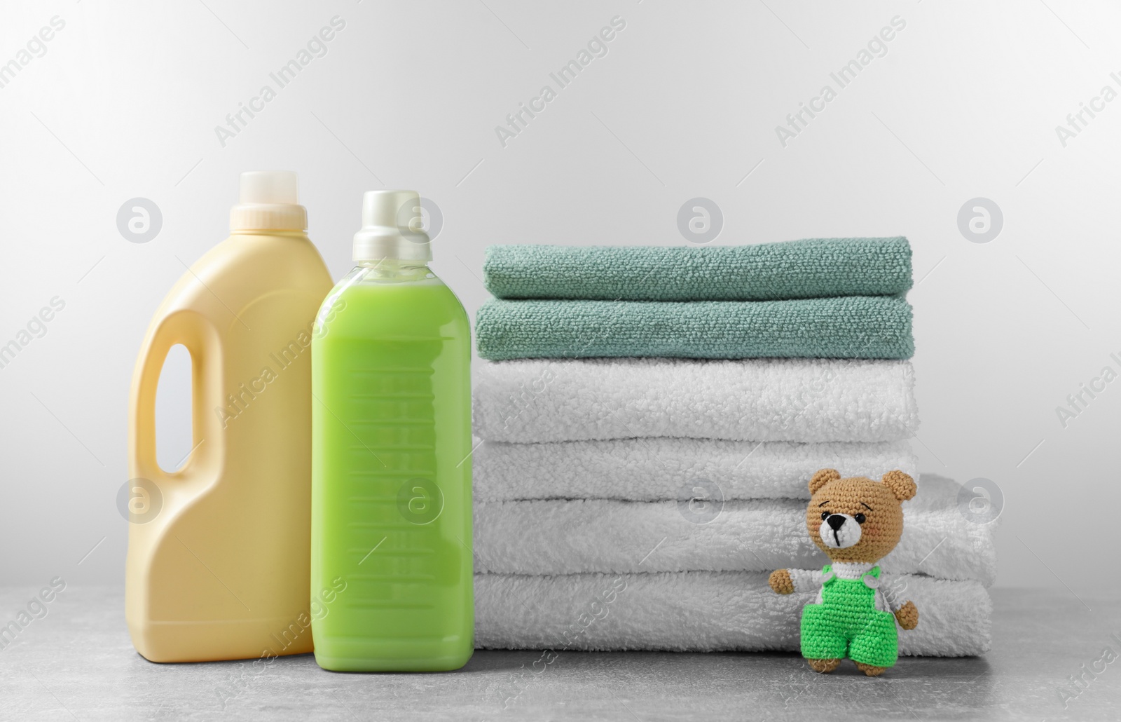 Photo of Bottles of laundry detergents, stacked fresh towels and knitted bear toy on table against white background