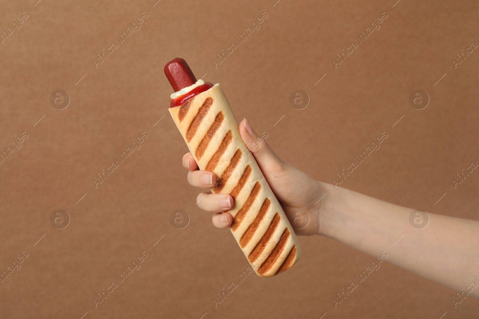 Photo of Woman holding delicious french hot dog on brown background, closeup