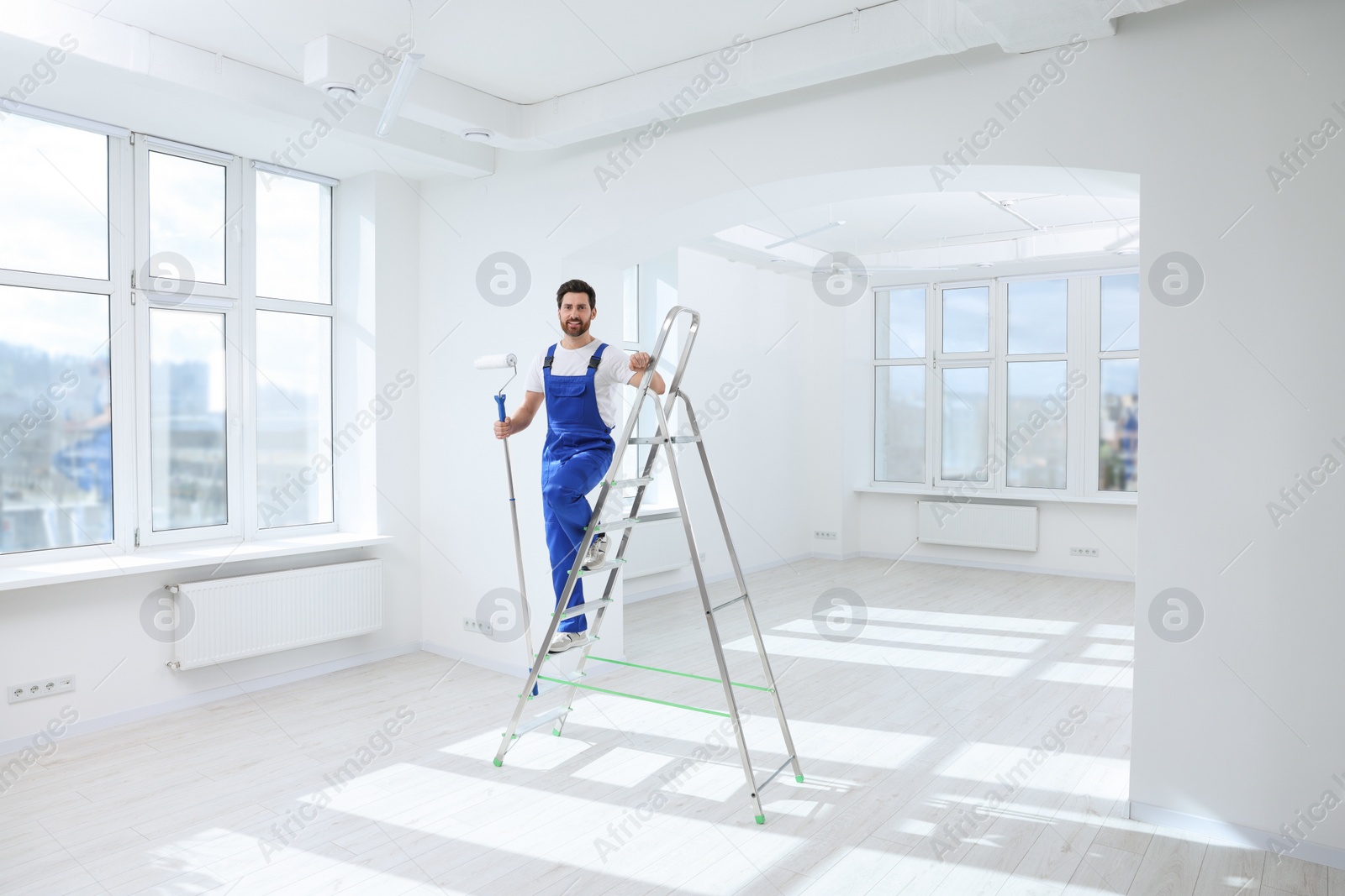 Photo of Handyman with roller on step ladder in room. Ceiling painting