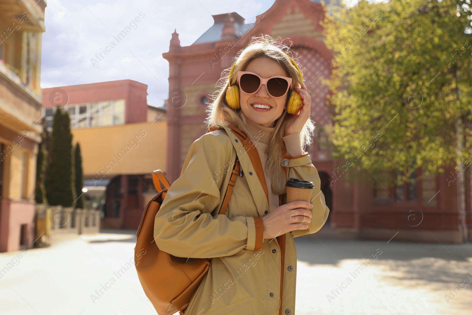 Photo of Happy young woman with coffee and headphones listening to music on city street