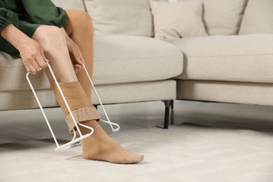 Woman putting on compression garment with stocking donner in living room, closeup and space for text. Prevention of varicose veins