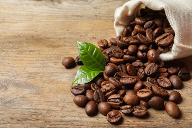Photo of Bag of coffee beans and fresh green leaves on wooden table, space for text