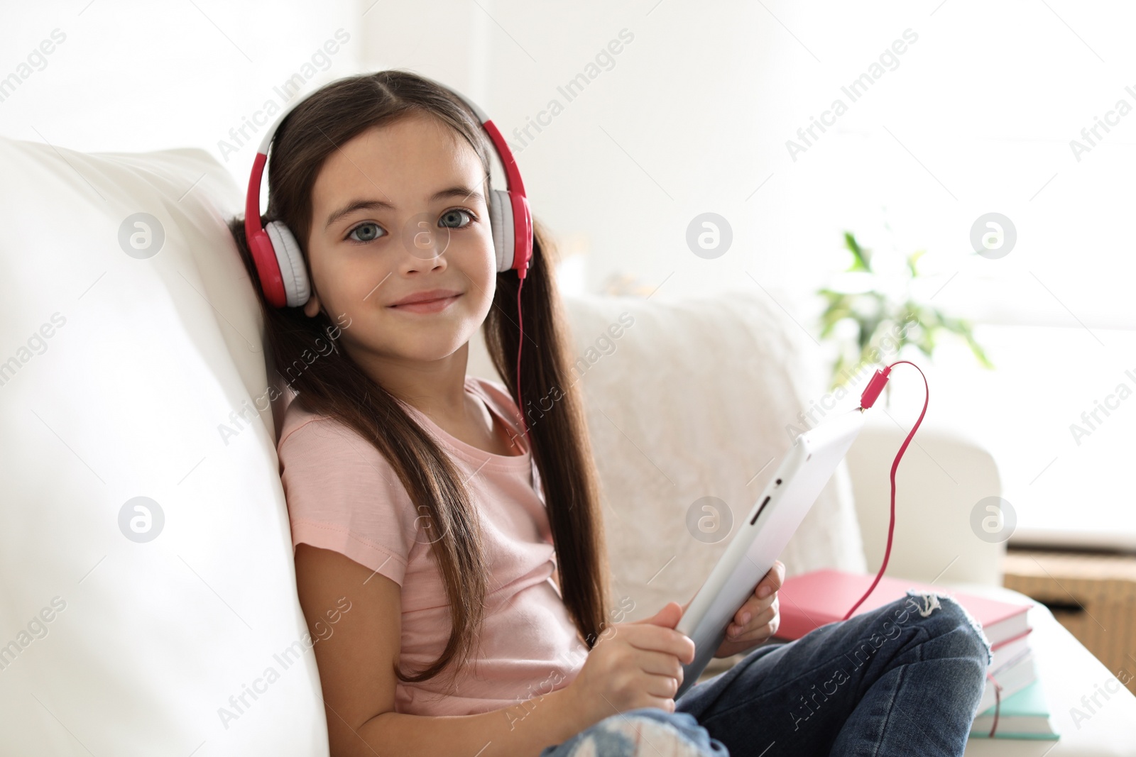 Photo of Cute little girl with headphones and tablet listening to audiobook at home