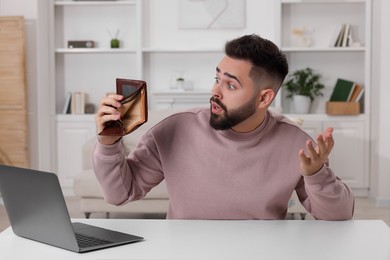 Confused man with empty wallet at white table indoors