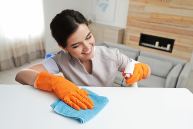 Photo of Young maid cleaning furniture with rag in hotel room