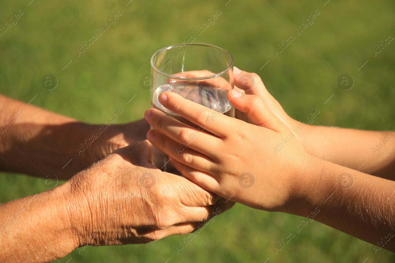 Photo of Child giving glass of water to elderly woman outdoors on sunny day, closeup