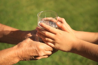 Photo of Child giving glass of water to elderly woman outdoors on sunny day, closeup