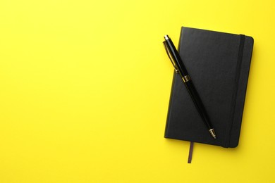 Closed black notebook and pen on yellow background, top view. Space for text