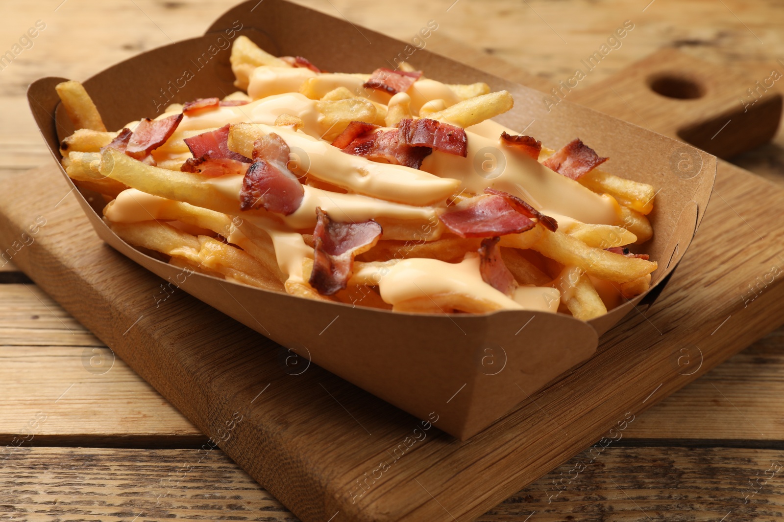 Photo of Tasty potato fries, cheese sauce and bacon in paper container on wooden table, closeup