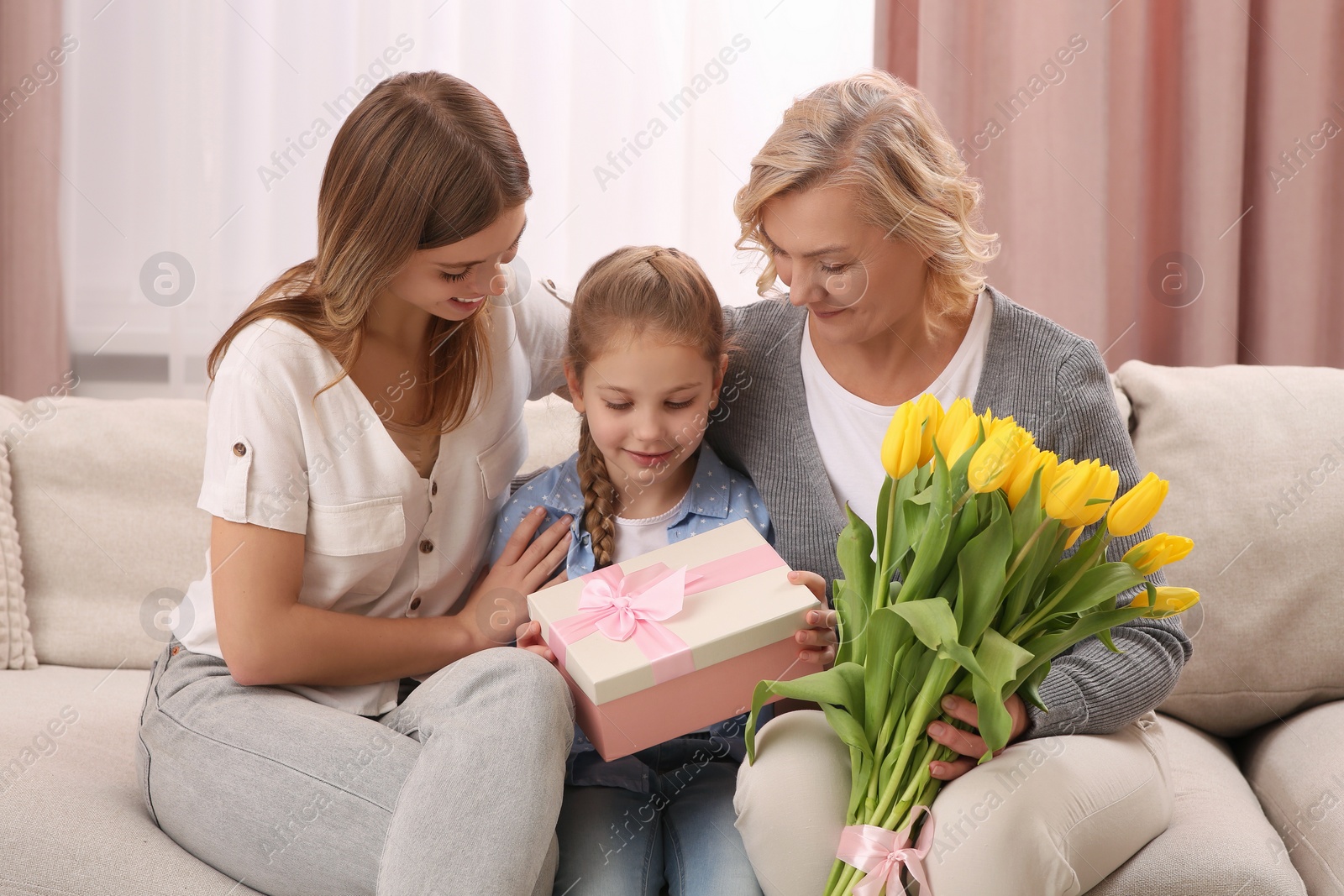 Photo of Little girl congratulating her mom and granny with flowers and gift at home. Happy Mother's Day