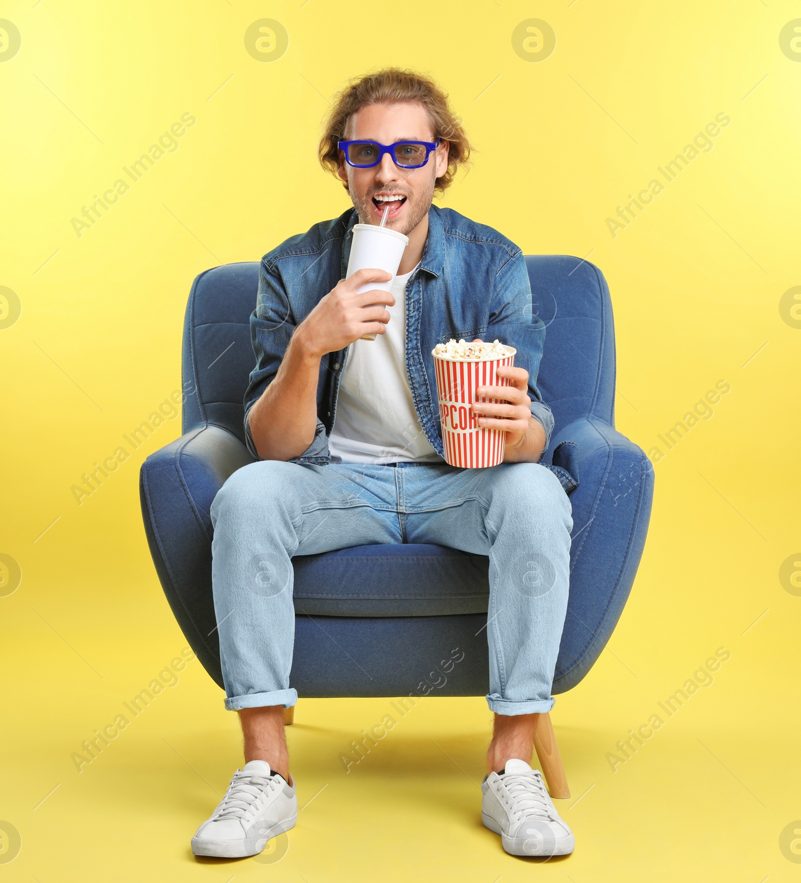 Photo of Man with 3D glasses, popcorn and beverage sitting in armchair during cinema show on color background