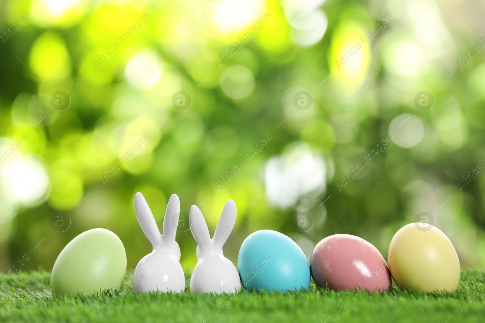 Photo of Ceramic Easter bunnies and dyed eggs on green grass against blurred background. Space for text