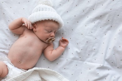 Cute newborn baby in white knitted hat sleeping on bed, top view