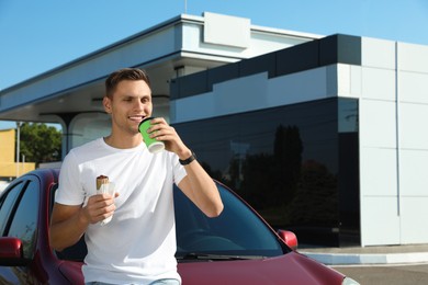 Photo of Young man with hot dog drinking coffee near car at gas station