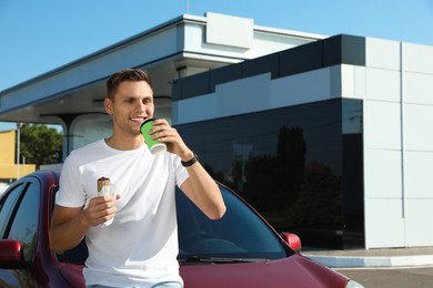 Young man with hot dog drinking coffee near car at gas station