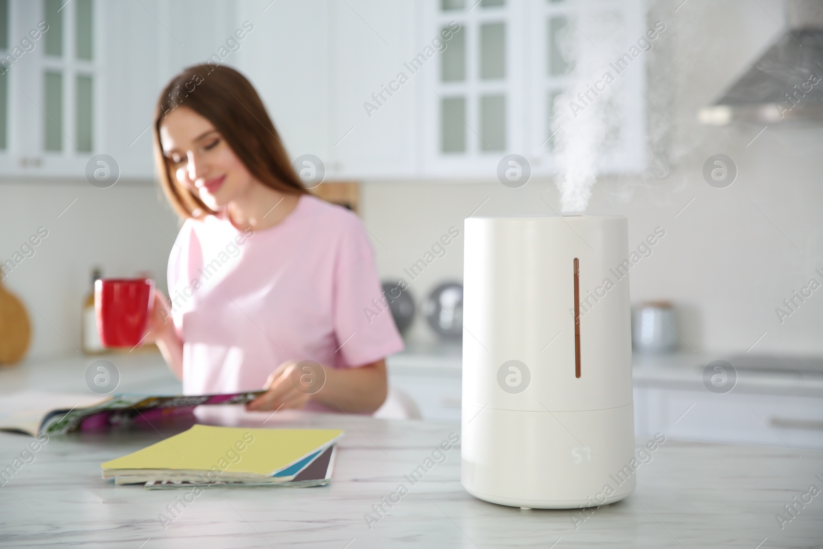 Photo of Modern air humidifier and blurred woman drinking coffee in kitchen