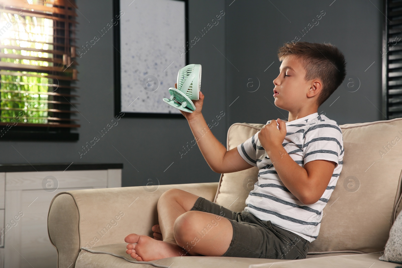 Photo of Little boy with portable fan suffering from heat at home. Summer season