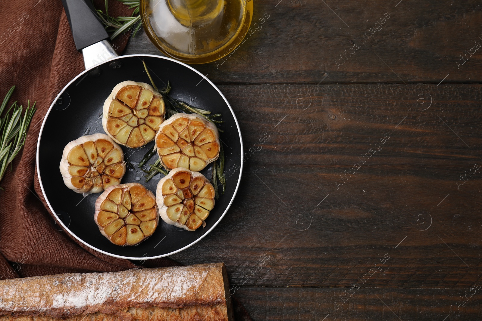 Photo of Frying pan with fried garlic, rosemary, oil and bread on wooden table, flat lay. Space for text