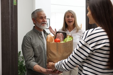 Photo of Courier giving paper bag with food products to senior couple indoors