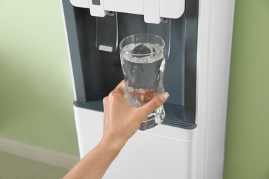 Woman filling glass from water cooler indoors, closeup. Refreshing drink