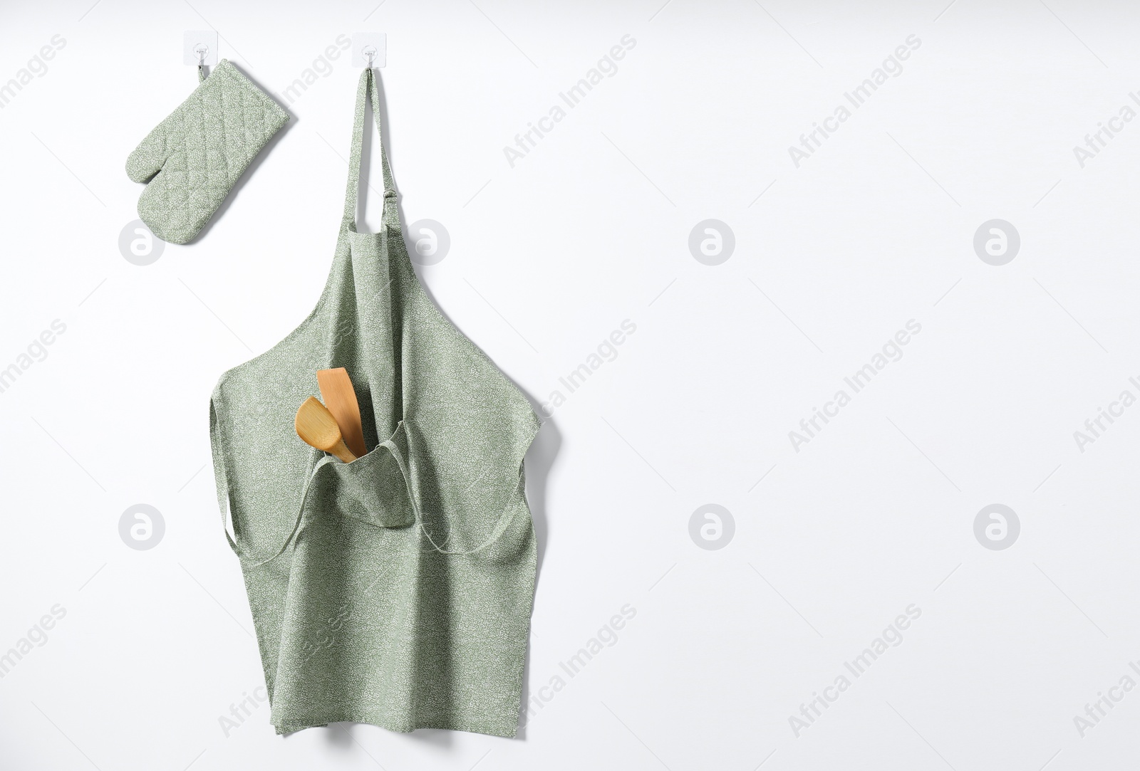 Photo of Clean apron with pattern, kitchen tools and oven glove on light wall. Space for text