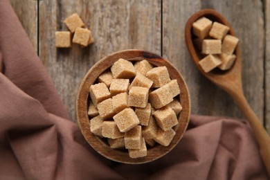 Photo of Bowl and spoon with brown sugar cubes on wooden table, flat lay