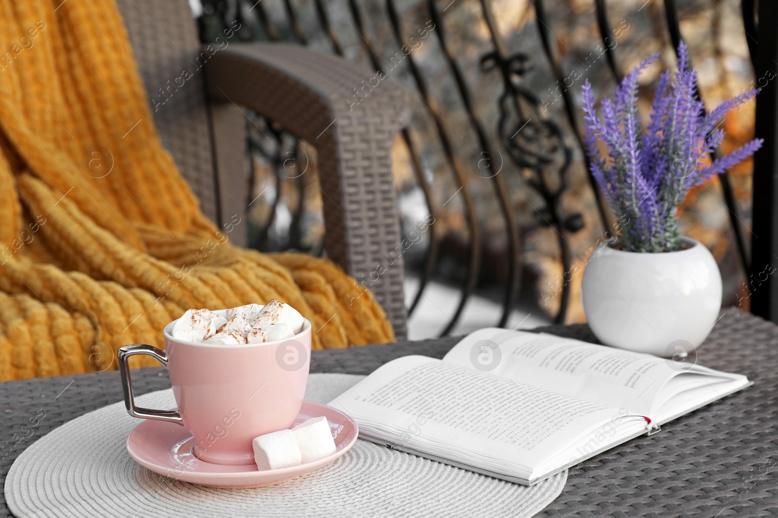 Photo of Cup of tasty cocoa with marshmallows and book on rattan table at balcony