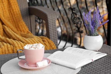 Photo of Cup of tasty cocoa with marshmallows and book on rattan table at balcony