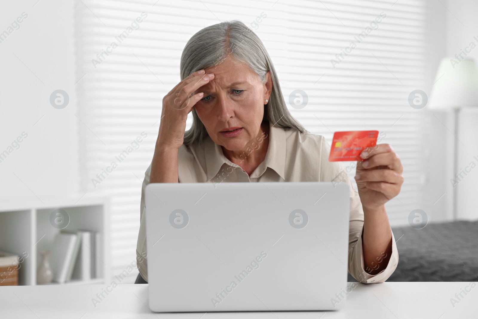 Photo of Upset woman with credit card near laptop at table indoors. Be careful - fraud