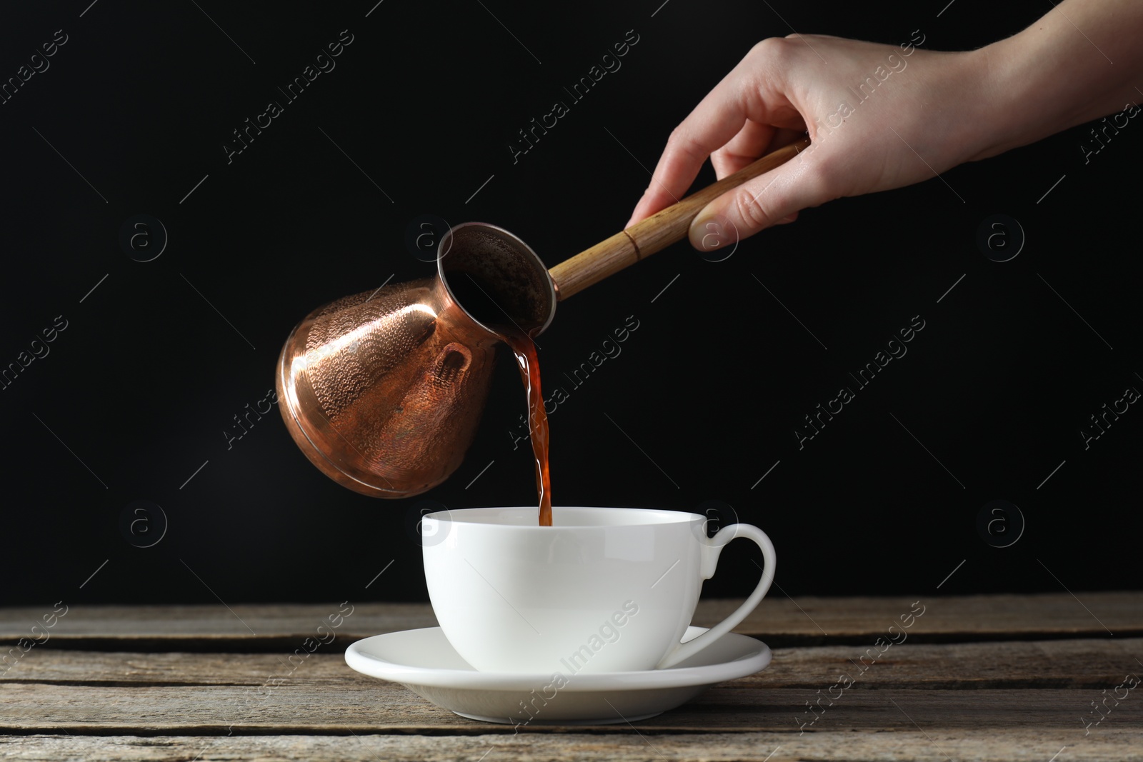 Photo of Turkish coffee. Woman pouring brewed beverage from cezve into cup at wooden table against black background, closeup