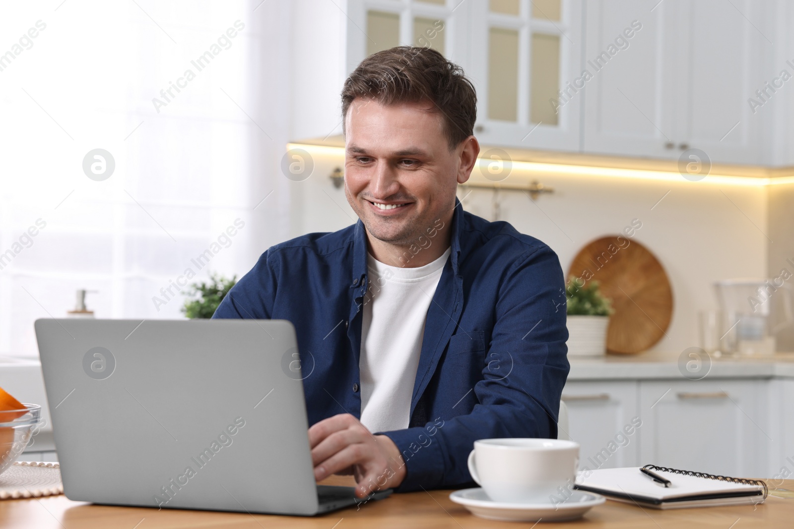 Photo of Happy man using laptop at wooden table in kitchen