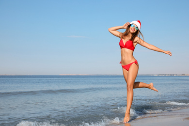 Photo of Young woman wearing Santa hat and bikini on beach, space for text. Christmas vacation