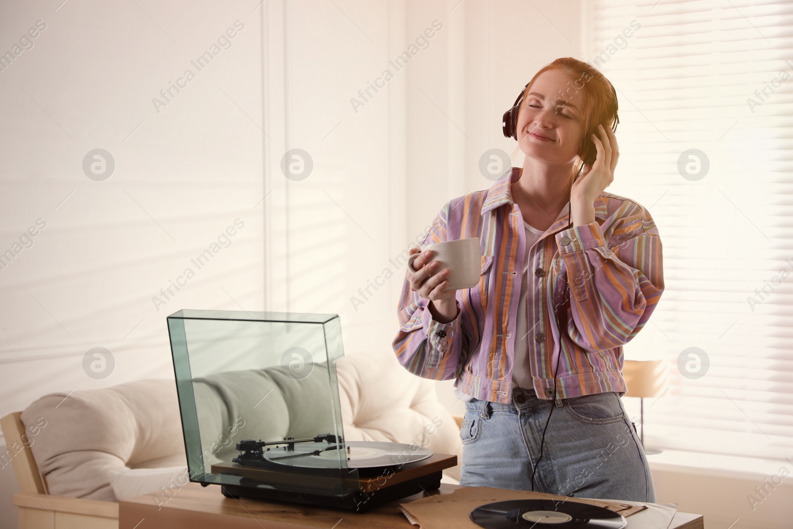 Photo of Young woman drinking coffee while listening to music with turntable in living room