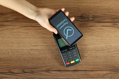 Woman using terminal for contactless payment with smartphone at wooden table, top view. Transaction completed screen on device