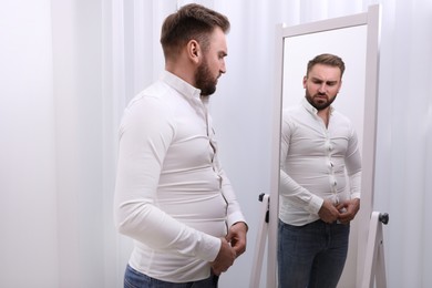 Photo of Upset man wearing tight shirt in front of mirror at home. Overweight problem