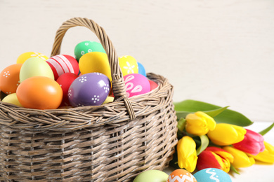 Photo of Colorful Easter eggs in basket and tulips on white wooden table, closeup