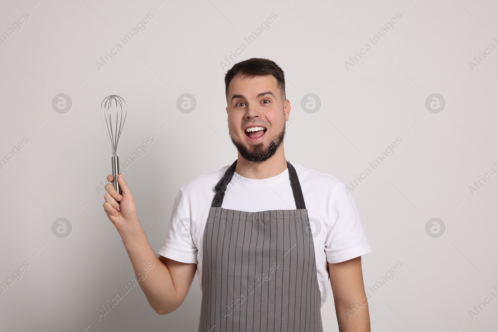 Photo of Excited professional confectioner in apron holding whisk on light grey background