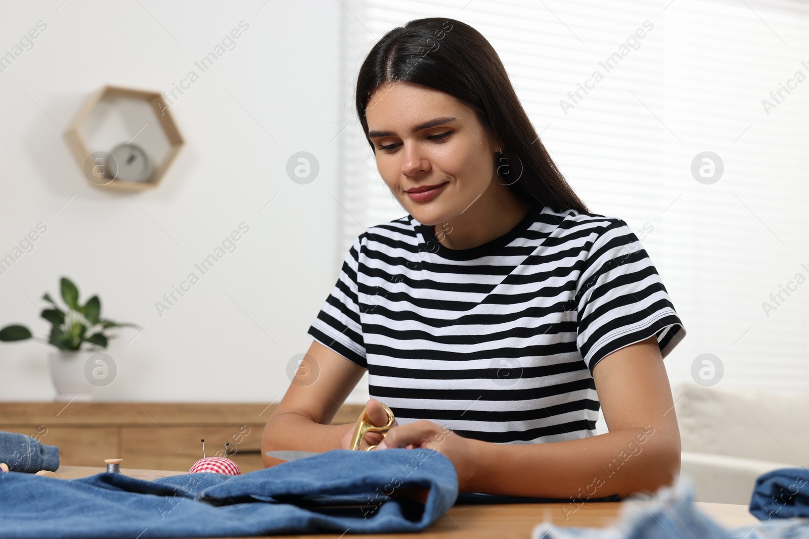 Photo of Young woman shortening jeans with scissors at wooden table indoors, space for text